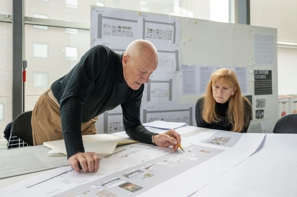 Arquitetos revisando projeto arquitetônico em mesa com plantas e painéis técnicos ao fundo.