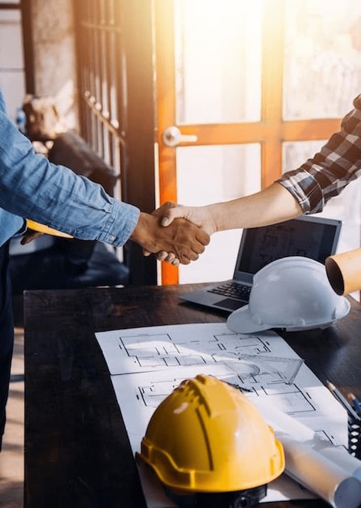 Aperto de mãos entre profissionais com capacetes e plantas sobre a mesa, representando parceria Prime com Arquiteto em projetos de arquitetura.